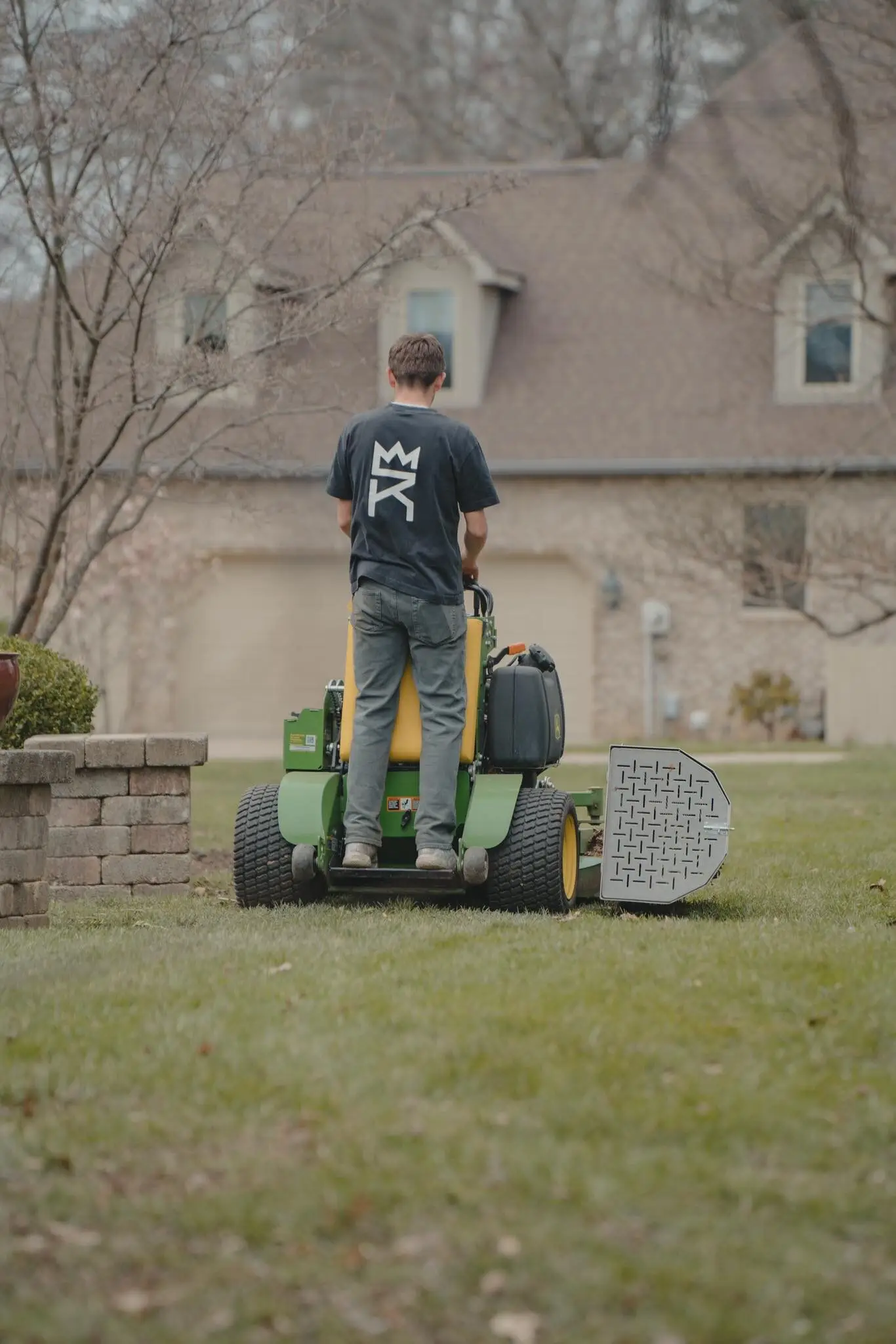 A crew member on a stand-on mower, facing a brick house across the lawn