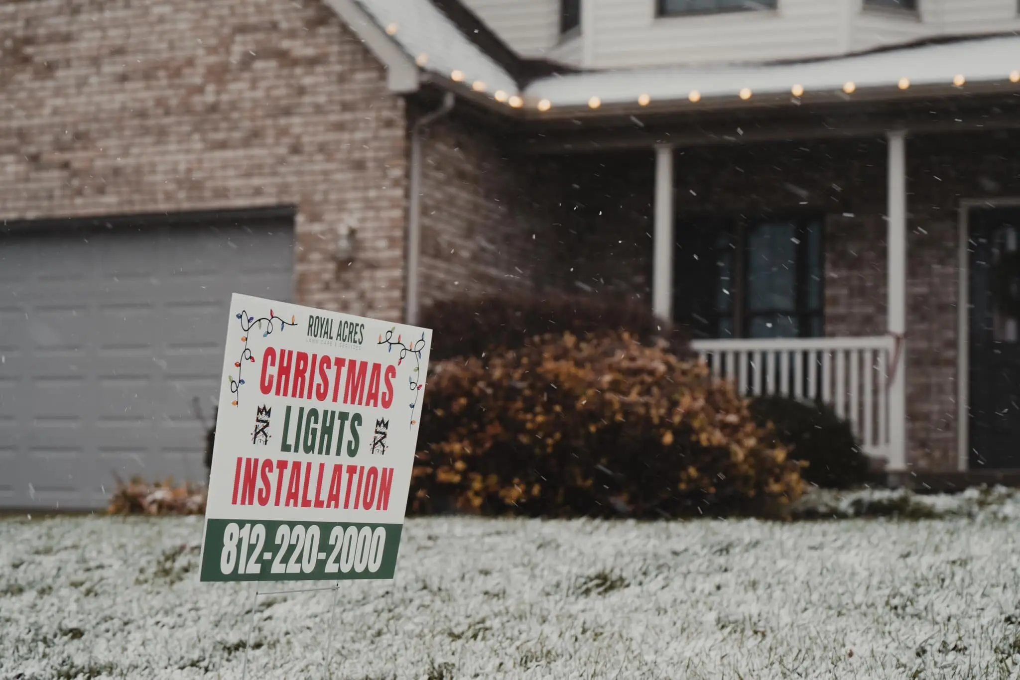 A Christmas lights installation sign on a frosted lawn, the house behind it lit