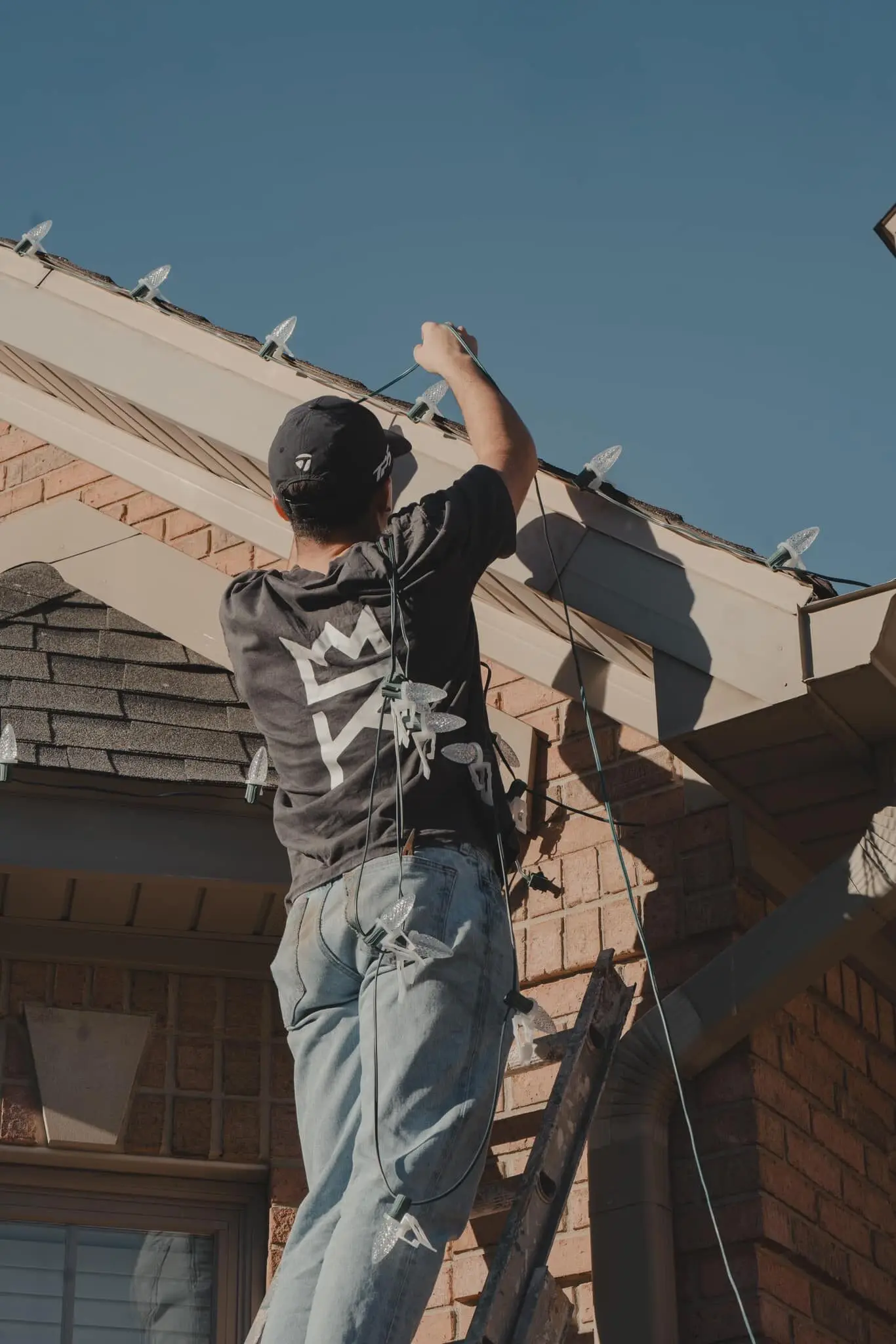 A crew member on a ladder clipping holiday lights to a roofline