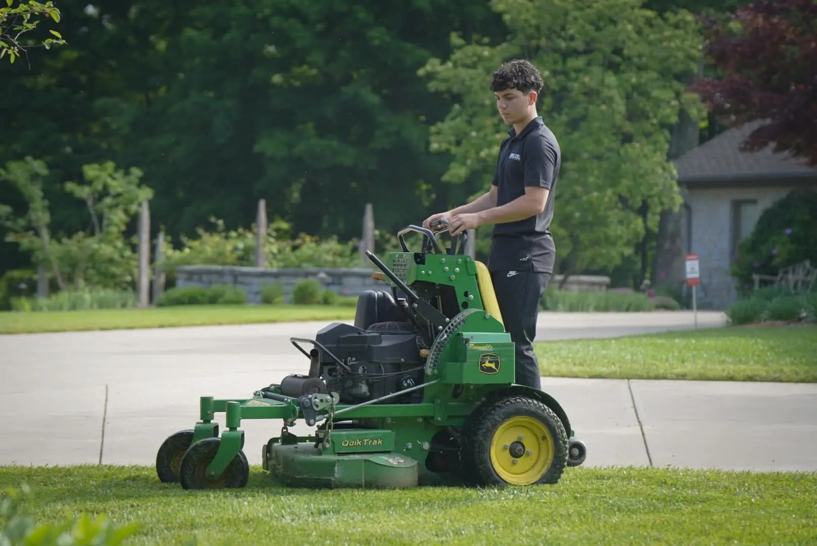 A crew member on a stand-on mower at the edge of a driveway