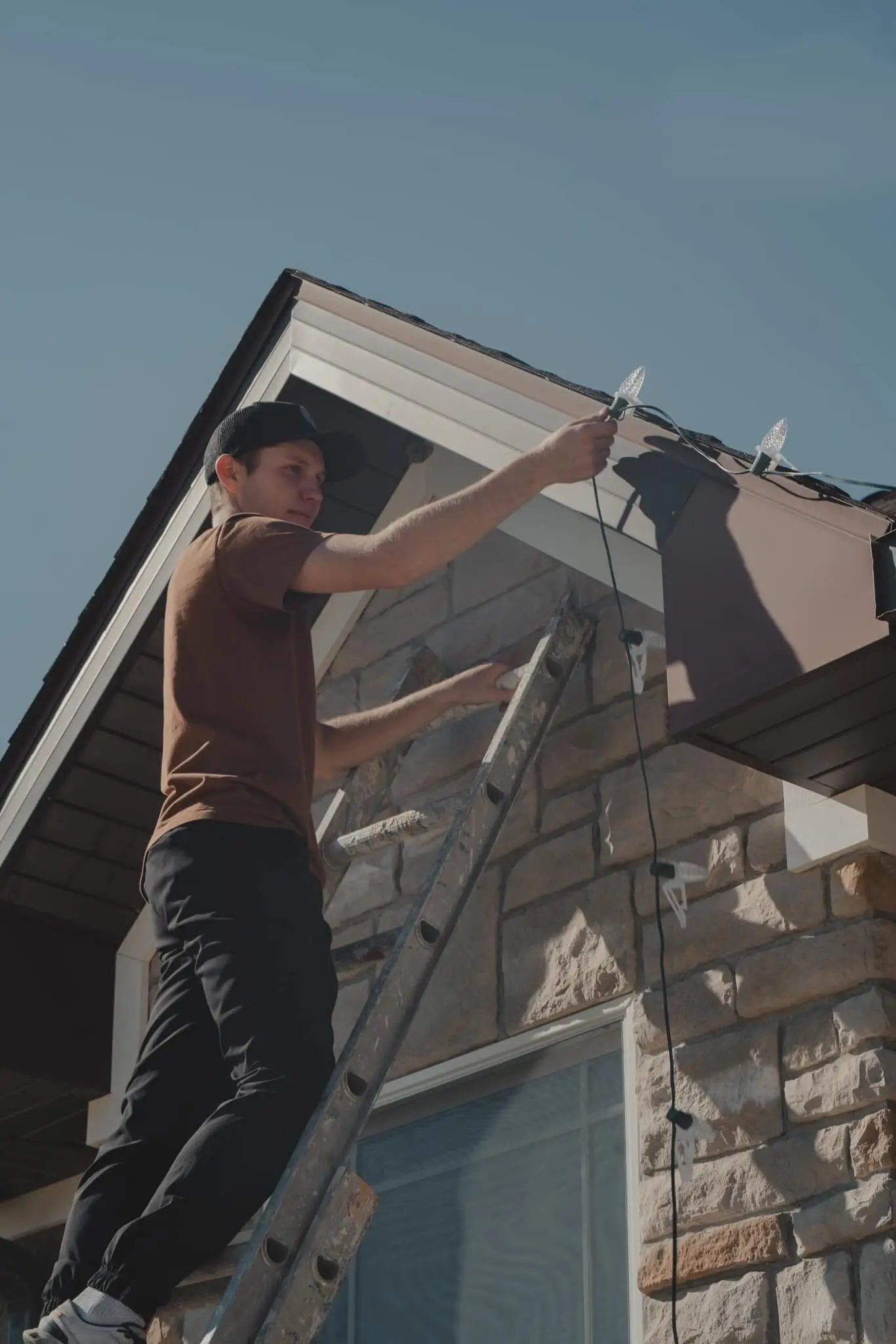 A crew member on a ladder hanging lights along a stone gable